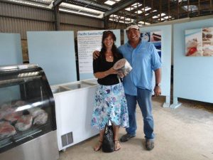 A woman and man stand smiling inside at Pacific Sunrise Fishing. The woman holds a large chunk of frozen tuna near a freezer that holds more tuna pieces.
