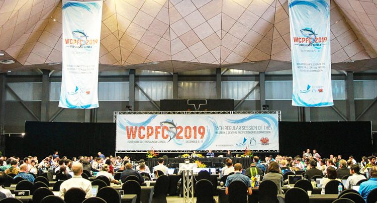 Large meeting hall hung with banners marked 'WVPFC 2019'. Four people sitting at a table on a dias, and scores of people sitting in seats around rows of long tables, at WCPFC16 in Port Moresby