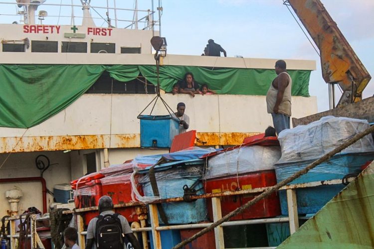 Part of small ship, with part of a crane visible, and one man using it to load a large plastic chiller box onto the ship. A woman and two children watch from the ship. One the wharf, a man stands on top of several other chiller boxes